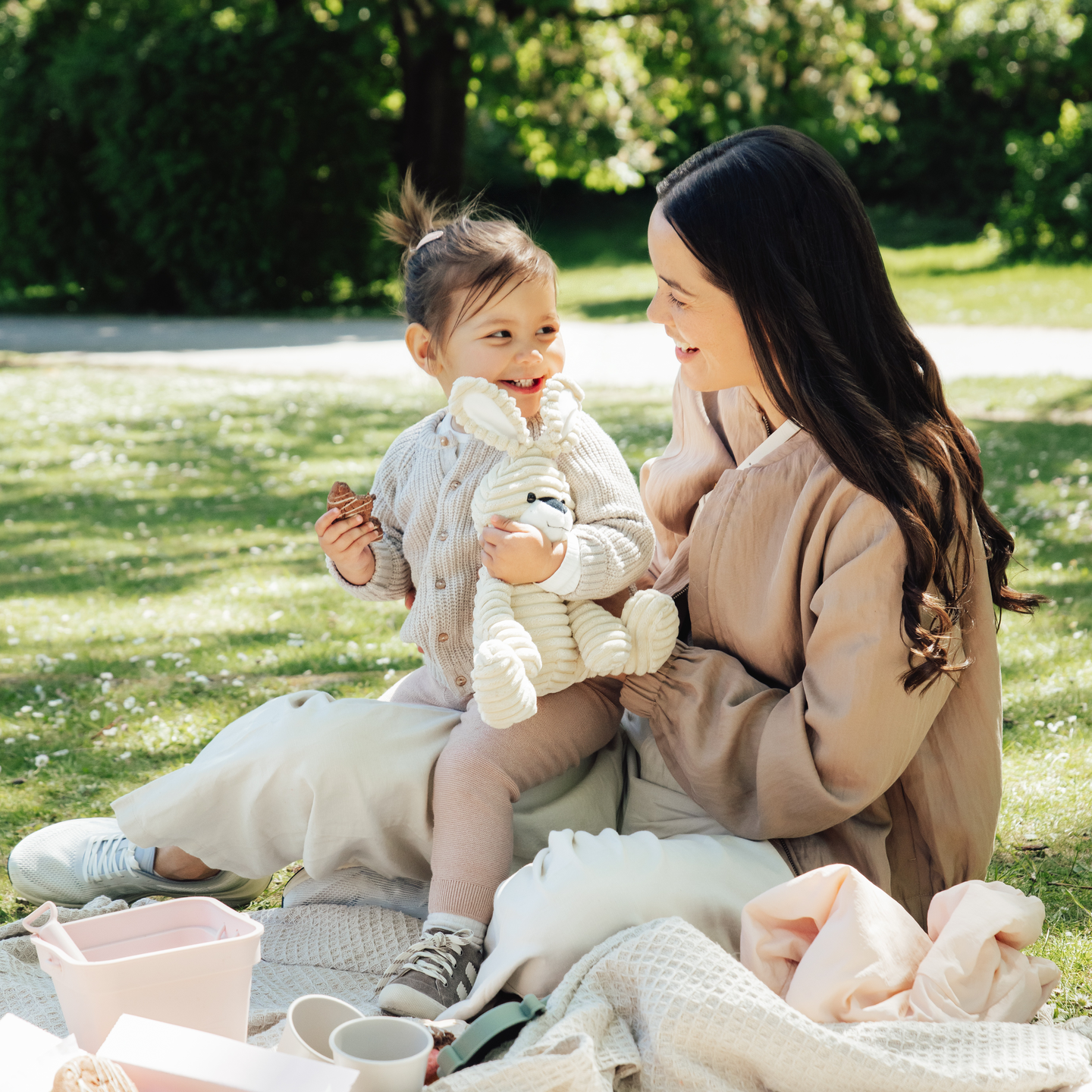 Une femme souriante est assise sur une couverture de pique-nique dans un parc, tenant un tout-petit qui rit et tient une peluche, avec la lumi&egrave;re du soleil filtrant &agrave; travers les arbres en arri&egrave;re-plan.